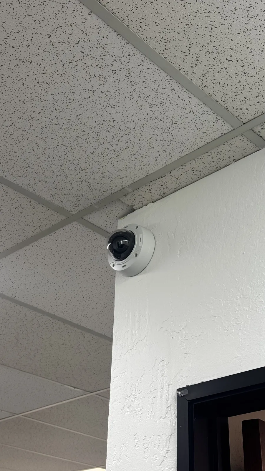 Dome camera mounted on the wall above a doorway at the edge of a drop ceiling
